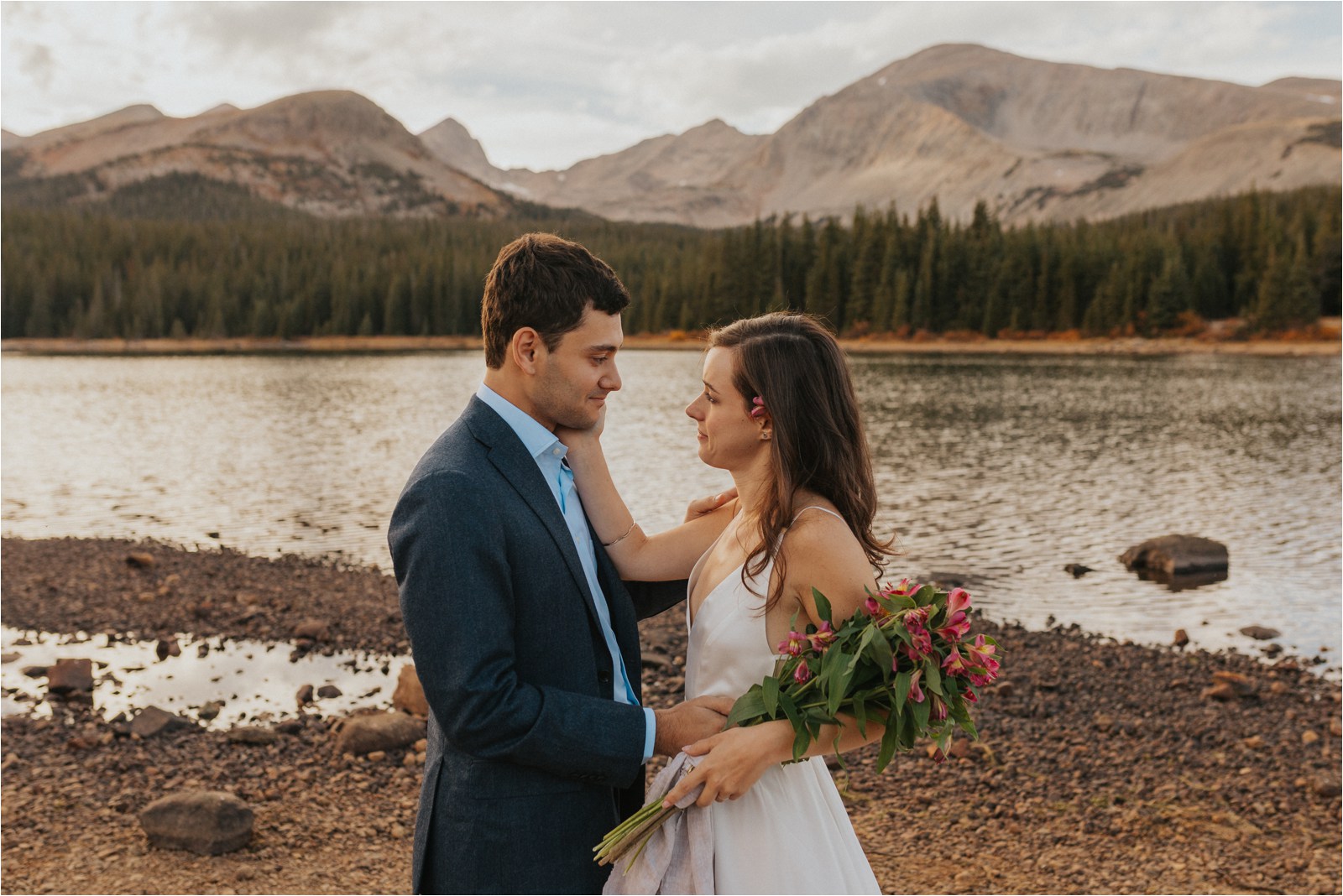 An emotional bride and groom embracing one another during their elopement in Colorado in front of a lake, with trees and snow-capped mountains in the distance.