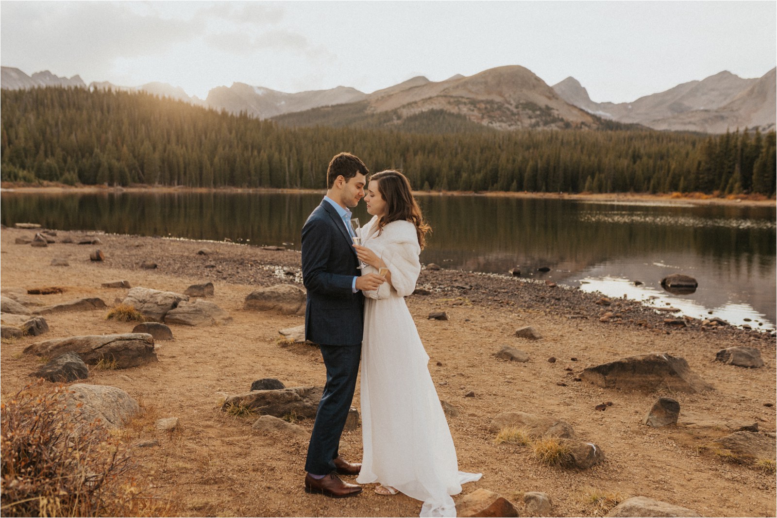 Married couple having their first dance and drinking champagne in front of a lake surrounded by mountains, with the sun setting between the mountain peaks after their elopement in Boulder, Colorado.