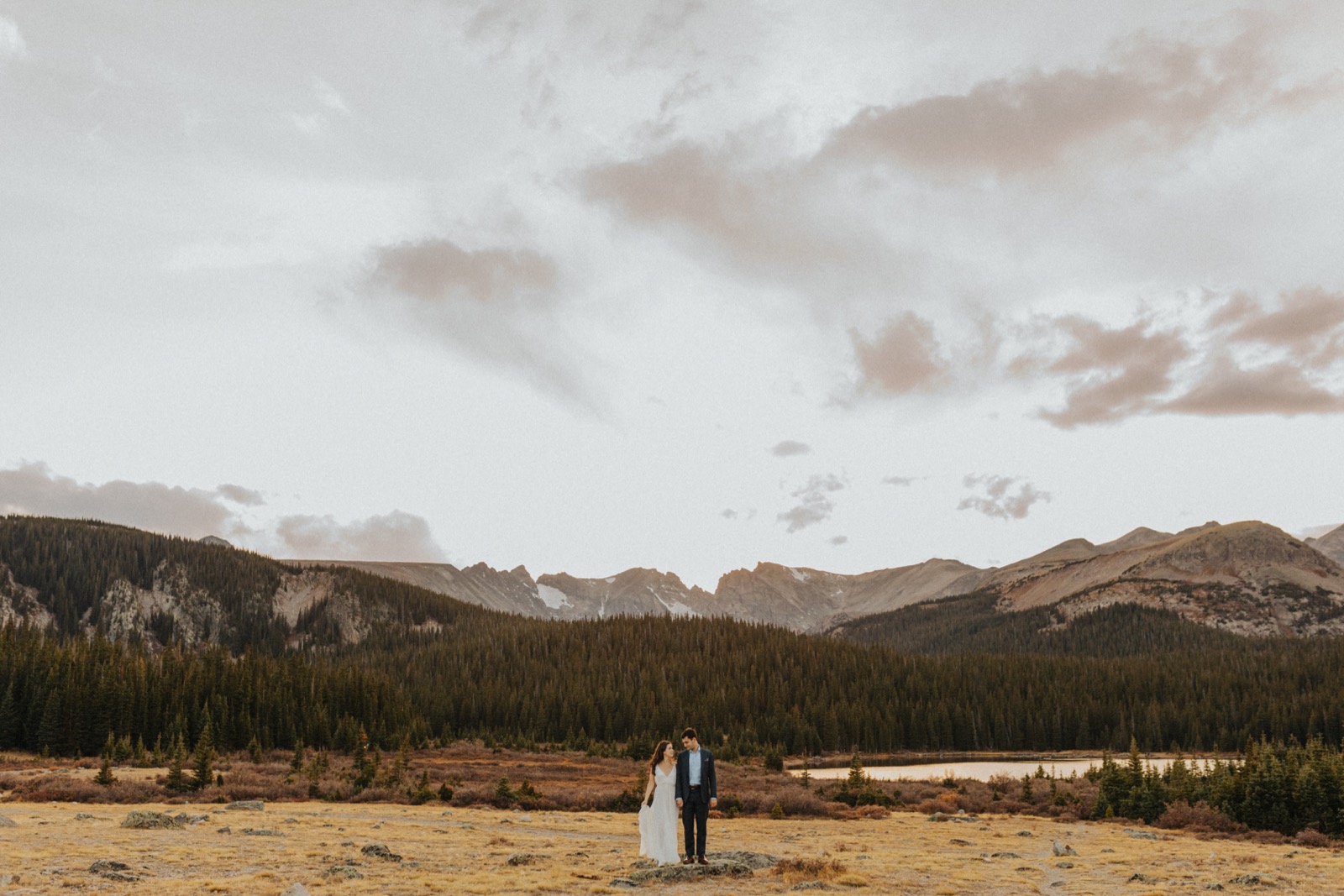 A couple that just had an elopement in Colorado standing in a rolling field, with the Rocky Mountains and a lake in the distance behind them.